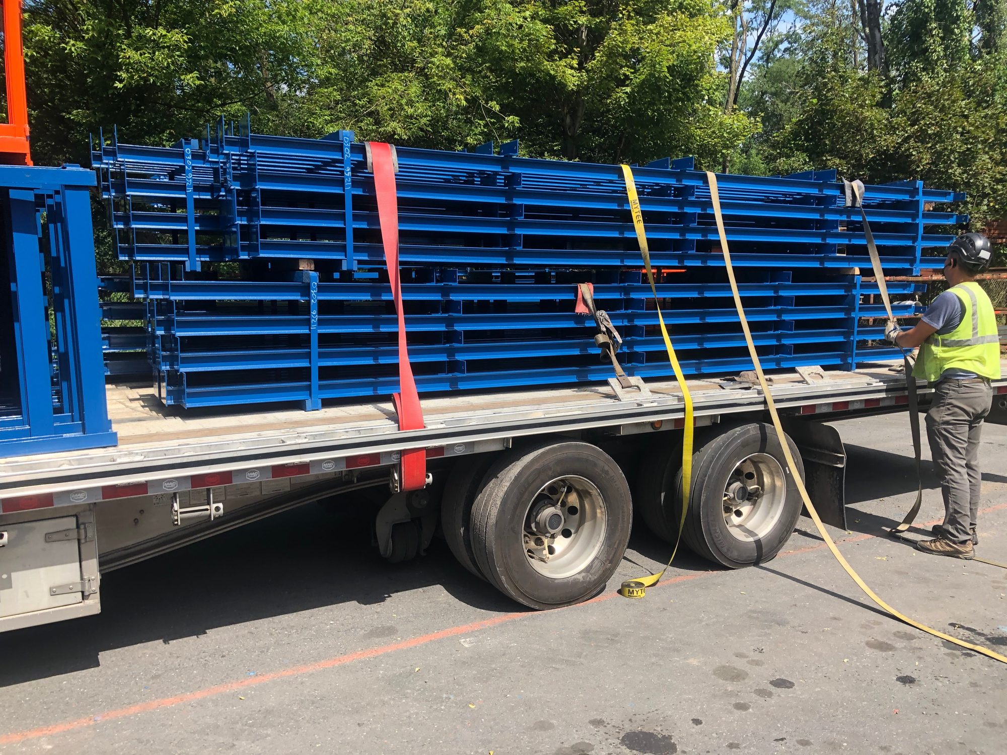A worker tensioning ratchet straps over stacked blue steel racking on a flatbed